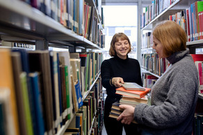 Zwei Studierende stehen zwischen zwei Bücherregalen mit Büchern in der Hand in der Bibliothek.