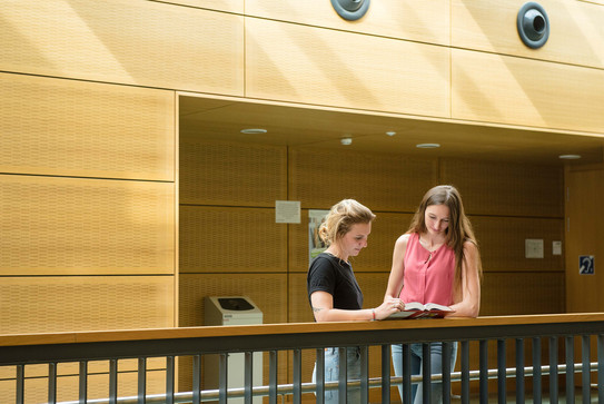 Zwei Studentinnen stehen mit einem Buch am Geländer.