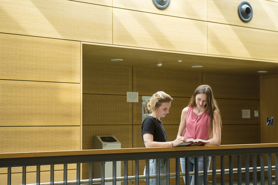 Zwei Studentinnen stehen mit einem Buch am Geländer.